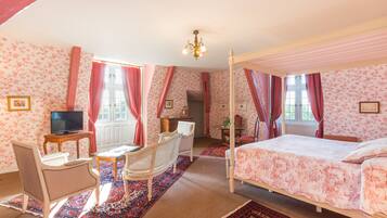 A four-poster bed with a floral bedspread, a television on a wooden stand, a wicker armchair, and a red and blue patterned rug.