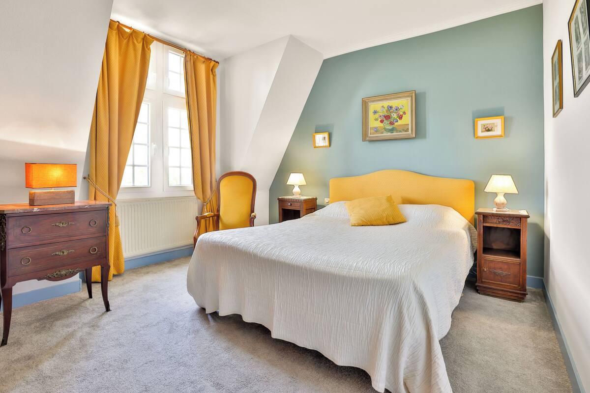 Une chambre à coucher comprenant un lit, deux tables de chevet, une chaise, une commode et une fenêtre avec des rideaux.