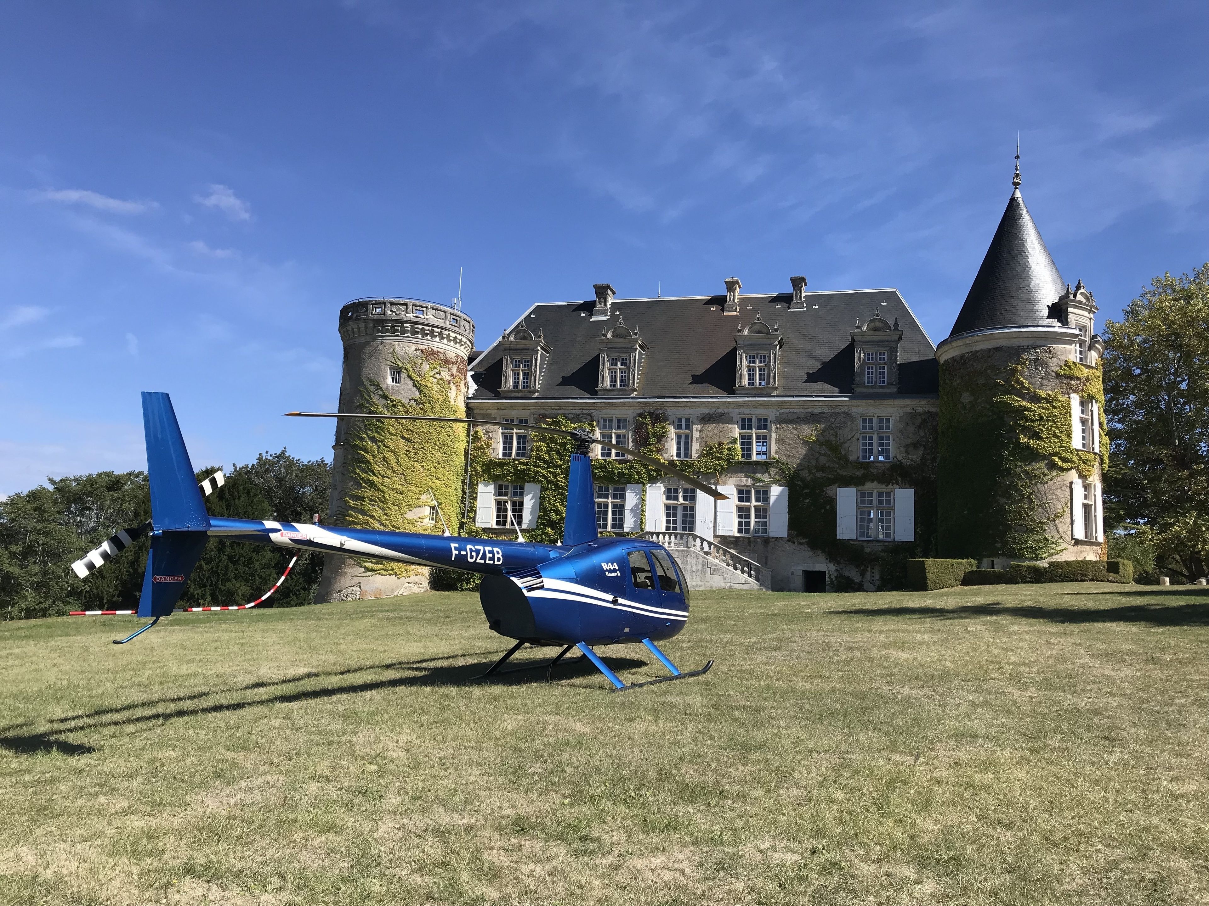 Un elicottero blu con targa F-GZEB parcheggiato su un prato di fronte a un grande edificio simile a un castello, con una torre e muri ricoperti d'edera.