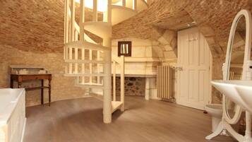 Una habitación con escalera de caracol, una bañera blanca, un espejo grande y una pared de piedra.