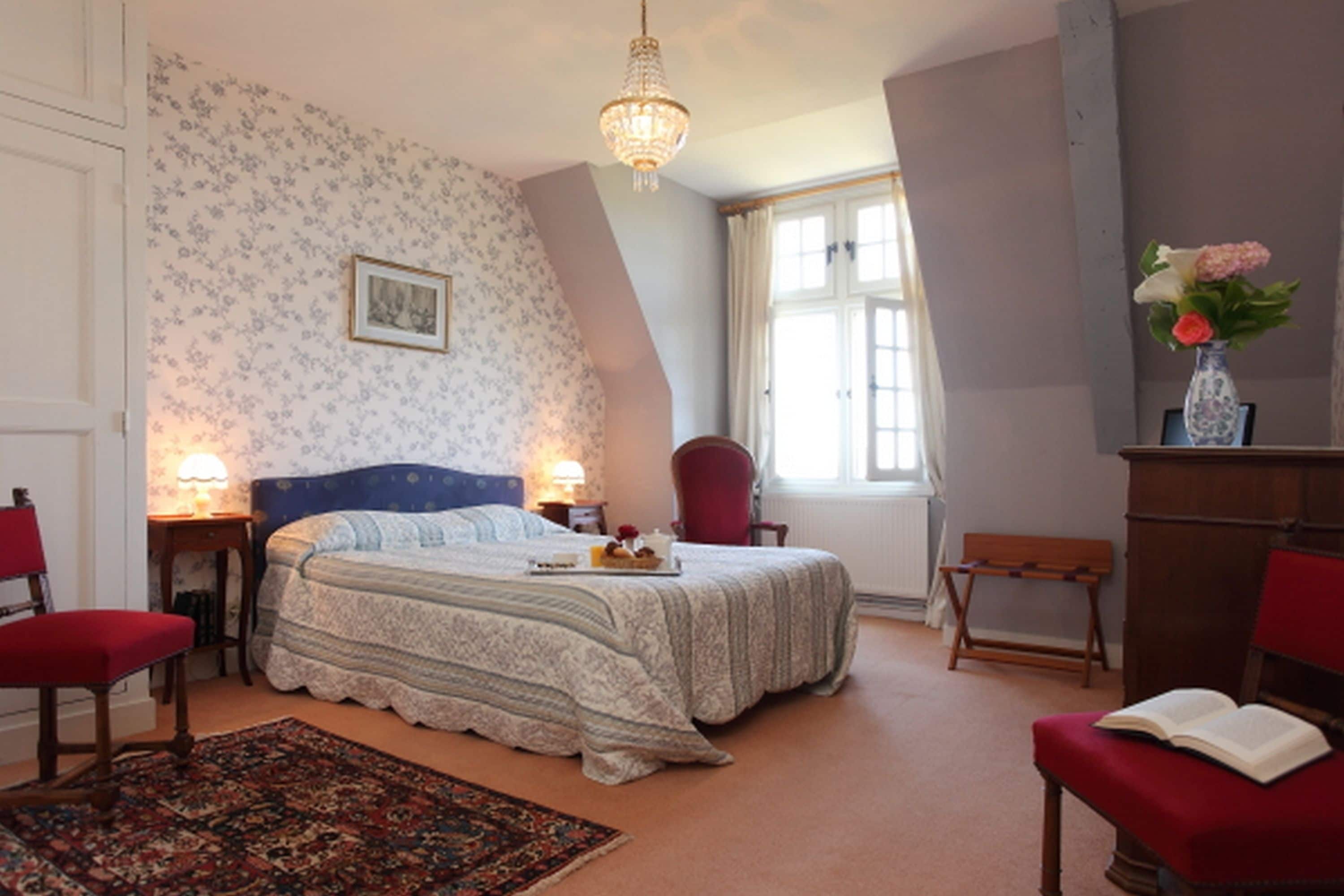 A bedroom with a bed, two bedside tables, a red chair, a wooden bench, a red ottoman, a wooden dresser, a chandelier, a window with curtains, and a floral wallpaper pattern.