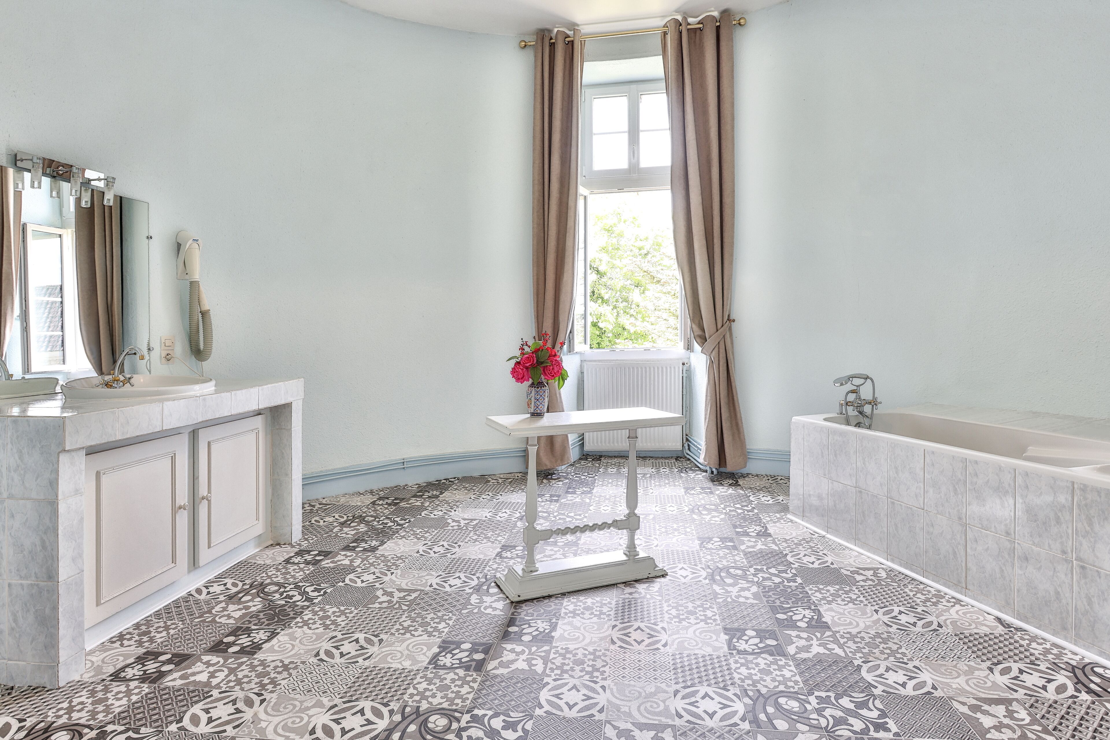 A bathroom with a patterned floor, a sink, a mirror, a vanity, a table with a vase of flowers, and a bathtub.
