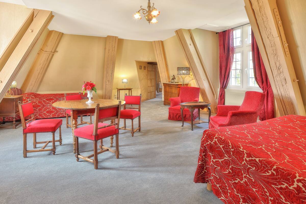 Une pièce avec un plafond haut, des poutres en bois, une table à manger avec des chaises, un lit recouvert d’une couverture rouge et une fenêtre avec des rideaux.