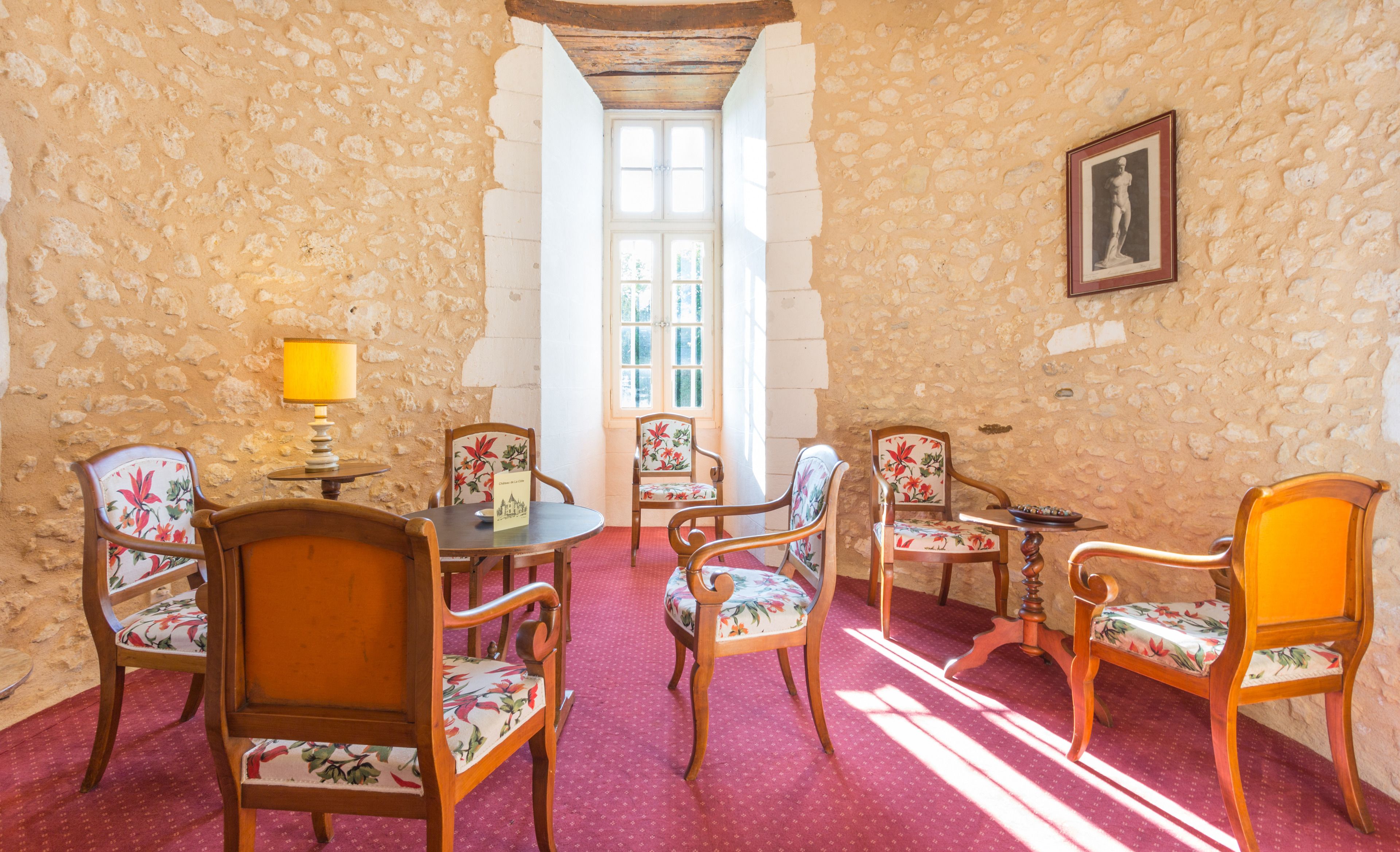 A room with stone walls, wooden furniture, and a red carpet. There is a window with white shutters, a table with a lamp, and chairs with floral upholstery.