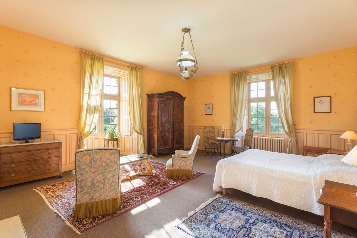 Une chambre à coucher comprenant un lit, deux chaises, une commode, un téléviseur et une fenêtre avec des rideaux.