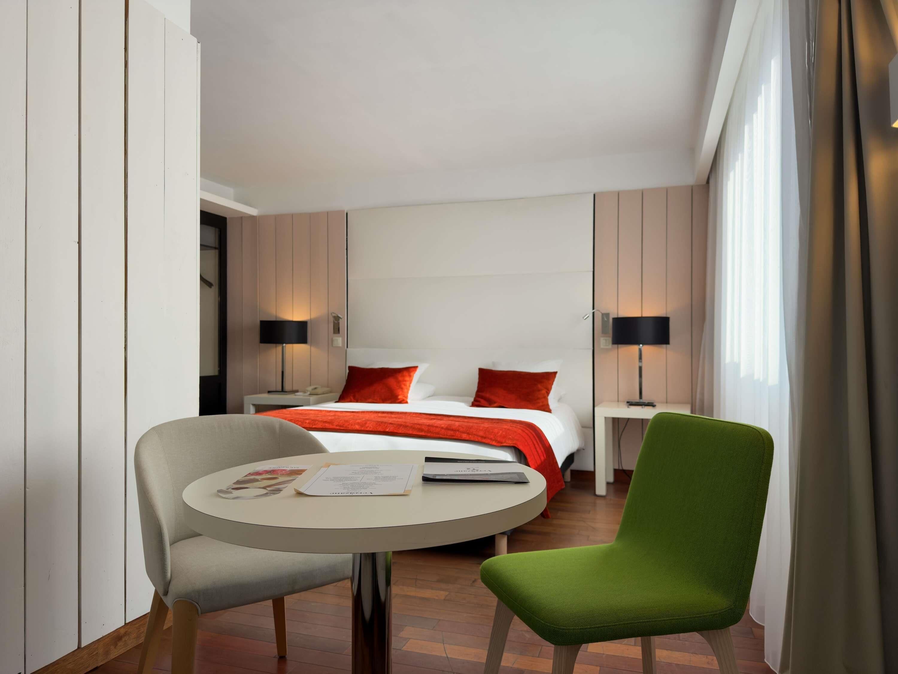 A hotel room with a bed, two bedside tables with lamps, a round table with a chair, and a window with curtains.