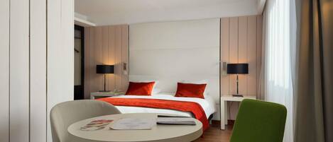 A hotel room with a bed, two bedside tables with lamps, a round table with a chair, and a window with curtains.
