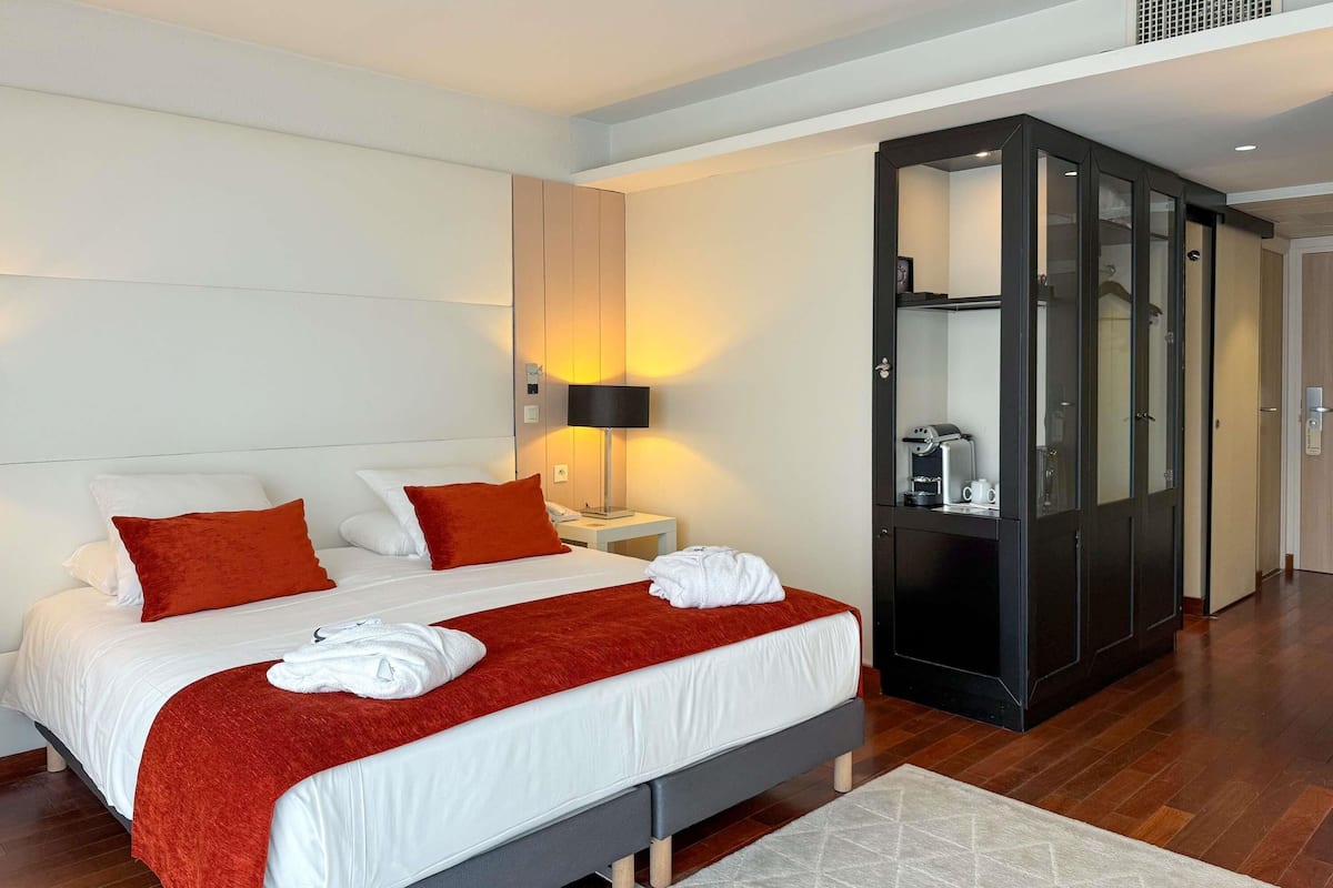 Una habitación de hotel moderna con una cama grande, un armario, una mesita de noche con lámpara y una cafetera.
