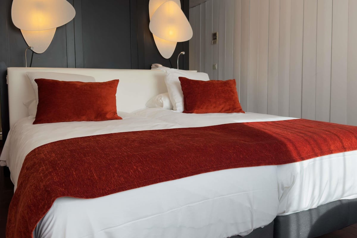 Una cama con ropa de cama blanca y roja, dos lámparas fijadas a la pared y un cabecero gris.