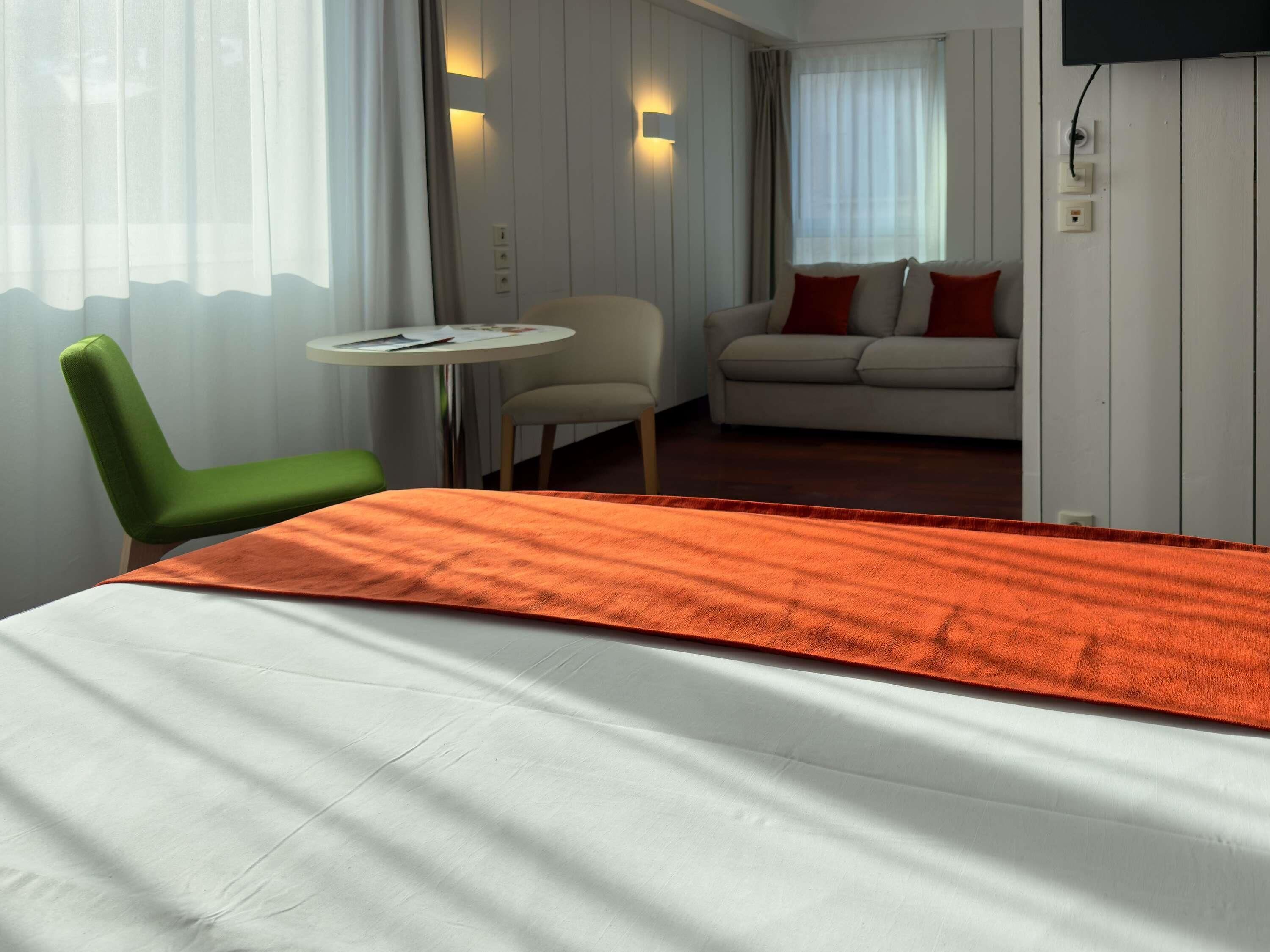 A hotel room with a bed, a green chair, a small round table, a sofa with red cushions, and a TV mounted on the wall.