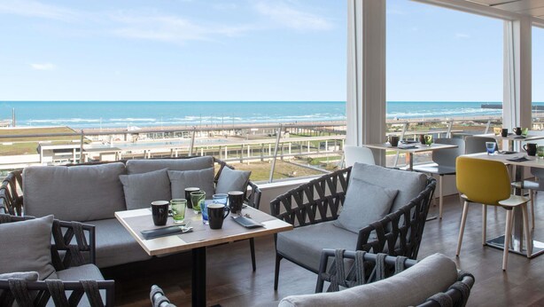 A modern dining area with a view of the ocean, featuring grey sofas, a wooden table, and various mugs and glasses. - Mercure Dieppe La Présidence (Dieppe)