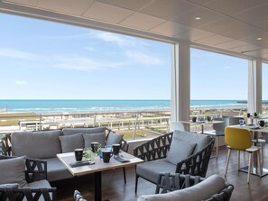 A modern dining area with a view of the ocean, featuring grey sofas, a wooden table, and various mugs and glasses. - Mercure Dieppe La Présidence (Dieppe)
