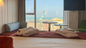 A hotel room with a bed, a window with a view of the beach and parking area, a small table, and a green chair. - Mercure Dieppe La Présidence (Dieppe)