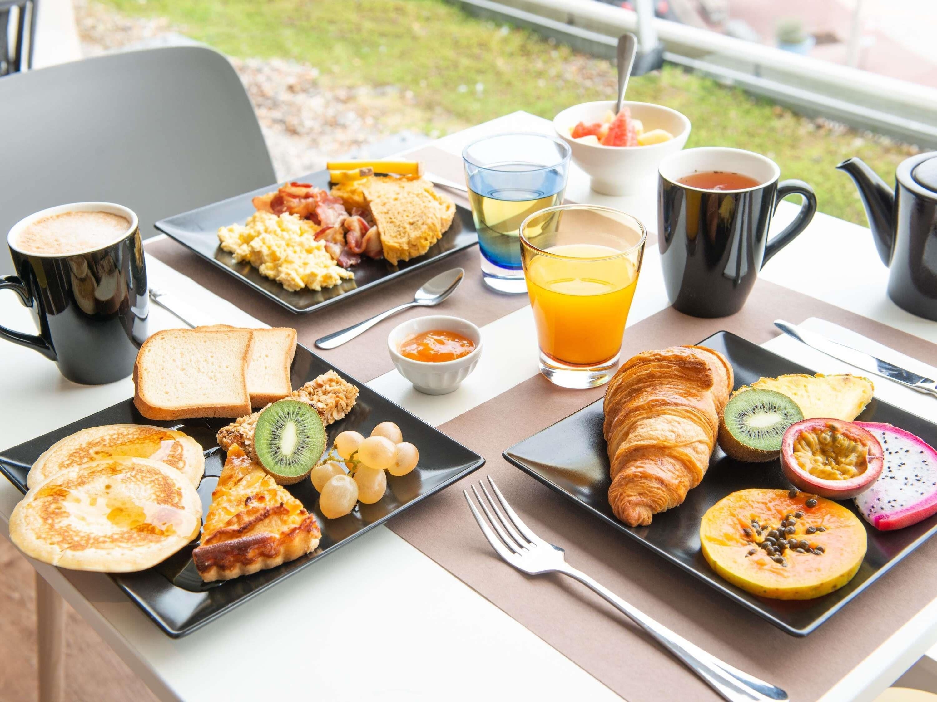 Una mesa de desayuno con tostadas, huevos, croissants y fruta. También hay tazas de café y jugo.
