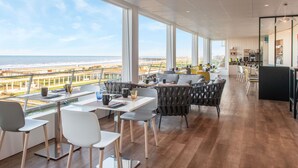 A modern dining area with a view of the beach, featuring a long table with chairs, a sofa, and a kitchen in the background. - Mercure Dieppe La Présidence (Dieppe)