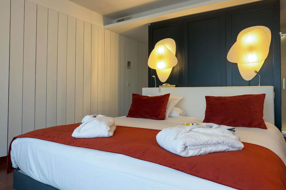 Habitación de hotel con una cama grande, dos almohadas rojas, un cabecero blanco y lámparas fijadas a la pared.