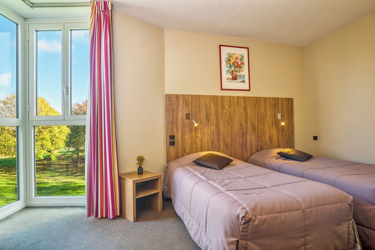 A bedroom with two beds, a wooden headboard, a small bedside table, a framed picture, and a view of a green landscape through large windows.