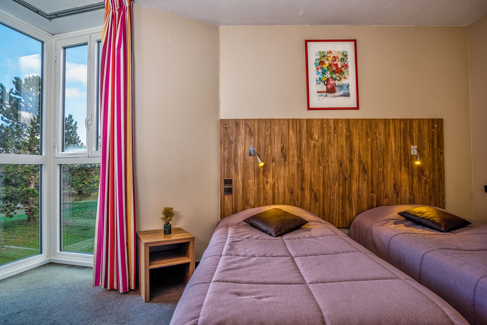 Habitación de hotel con dos camas, cabecero de madera, una mesita de noche con un florero y una ventana con cortinas.