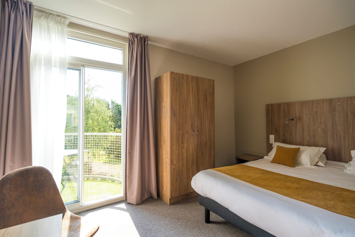 A hotel room with a bed, a wooden wardrobe, a chair, and a balcony with curtains.