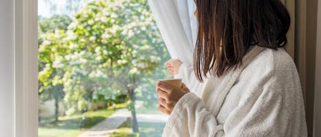 A person in a white robe holding a cup, standing by a window with a view of a garden and trees.