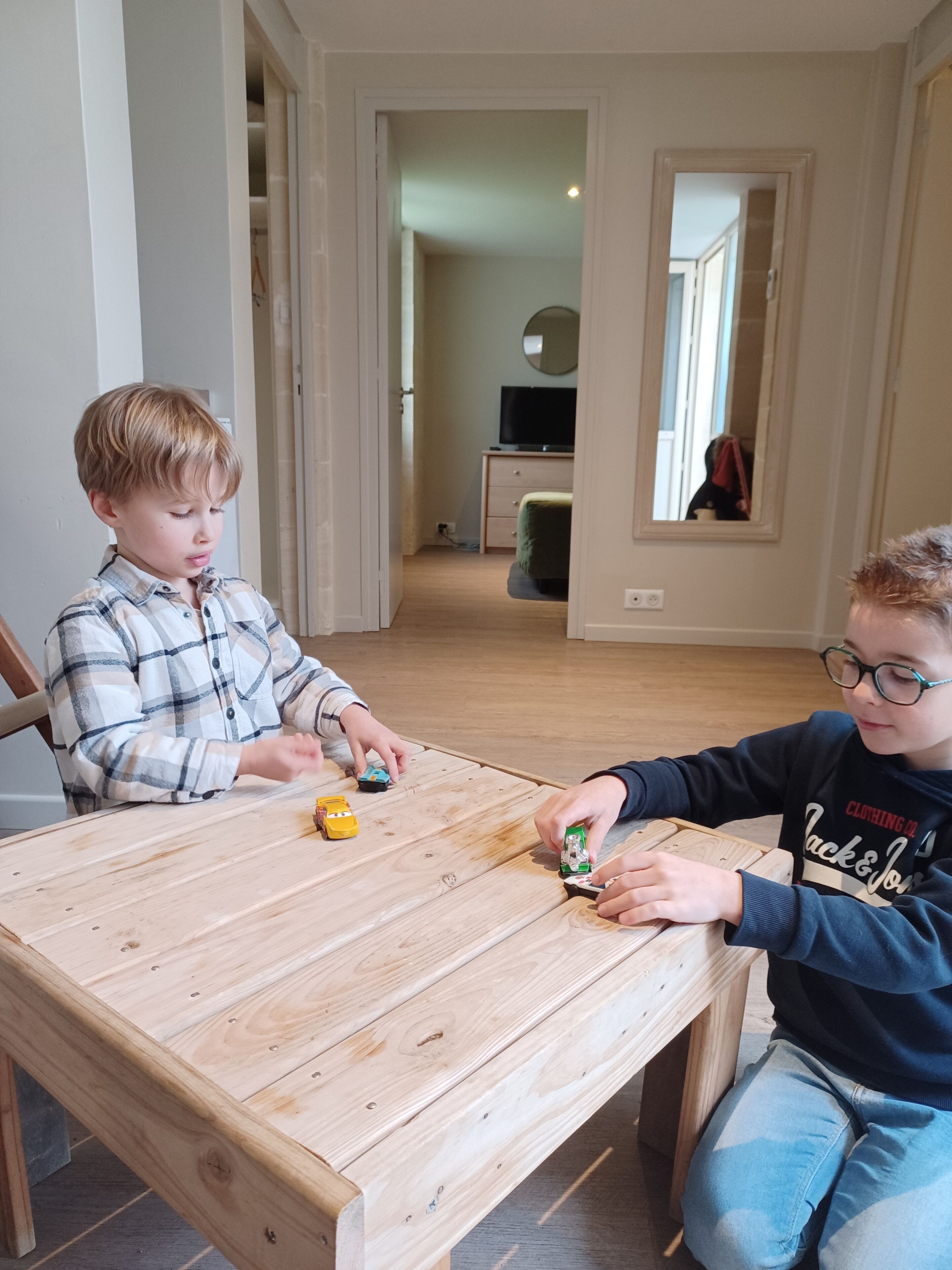 Two children playing with toy cars at a wooden table in a room with a mirror and a television in the background.