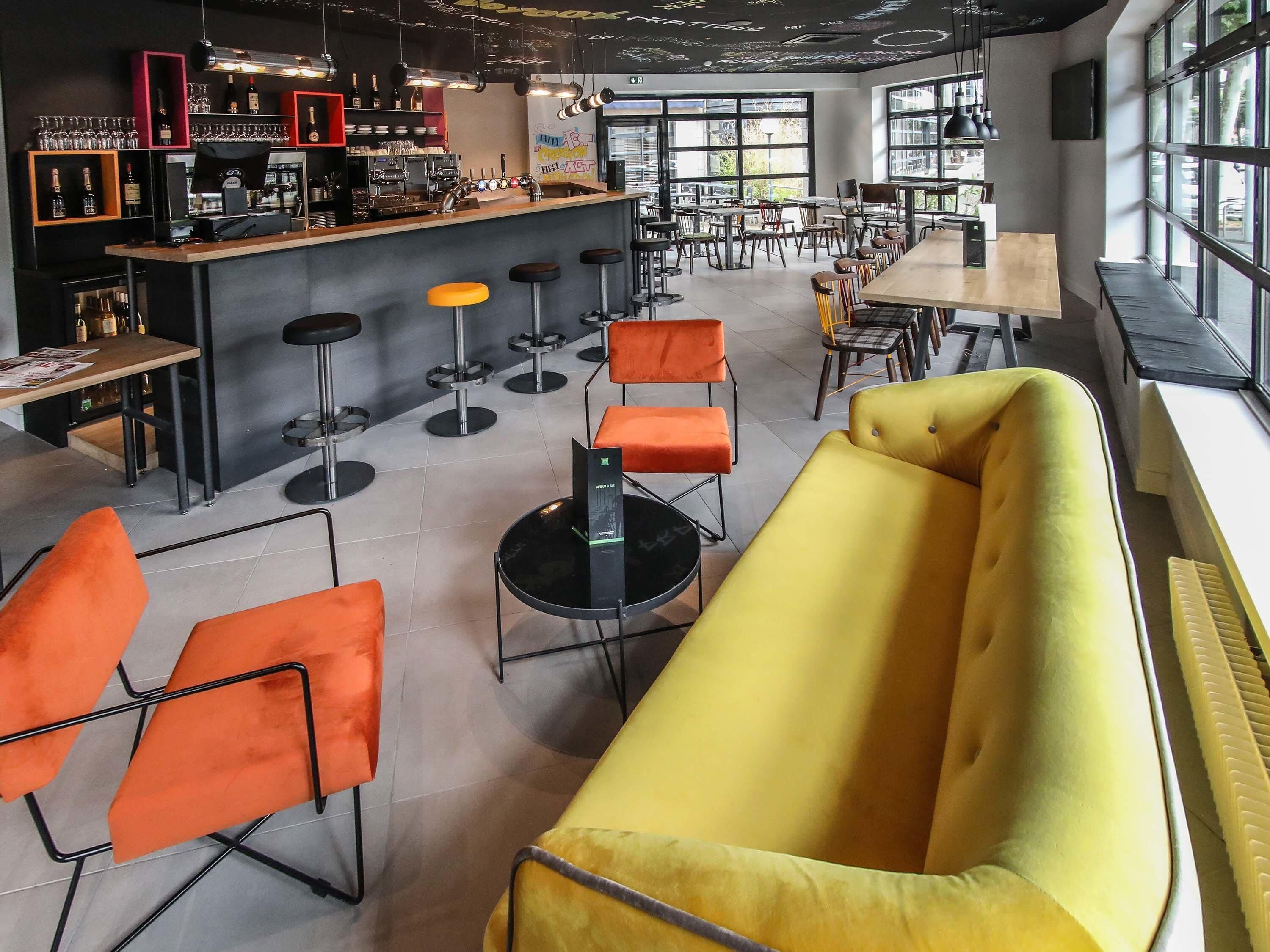 A modern bar area with stools, a counter, and a seating area with orange chairs and a yellow sofa.