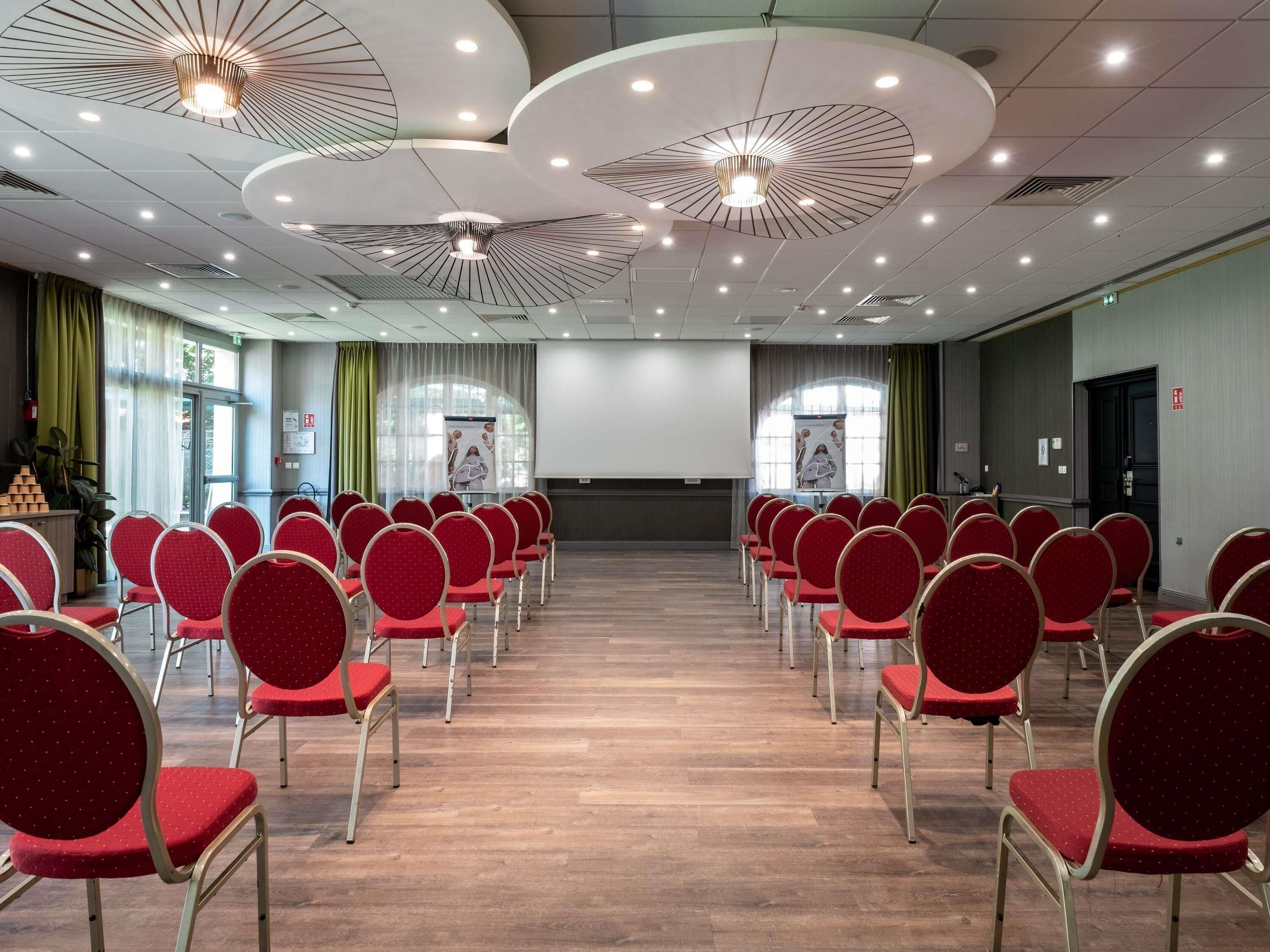 A conference room with rows of red chairs, a podium, and a large screen at the front.