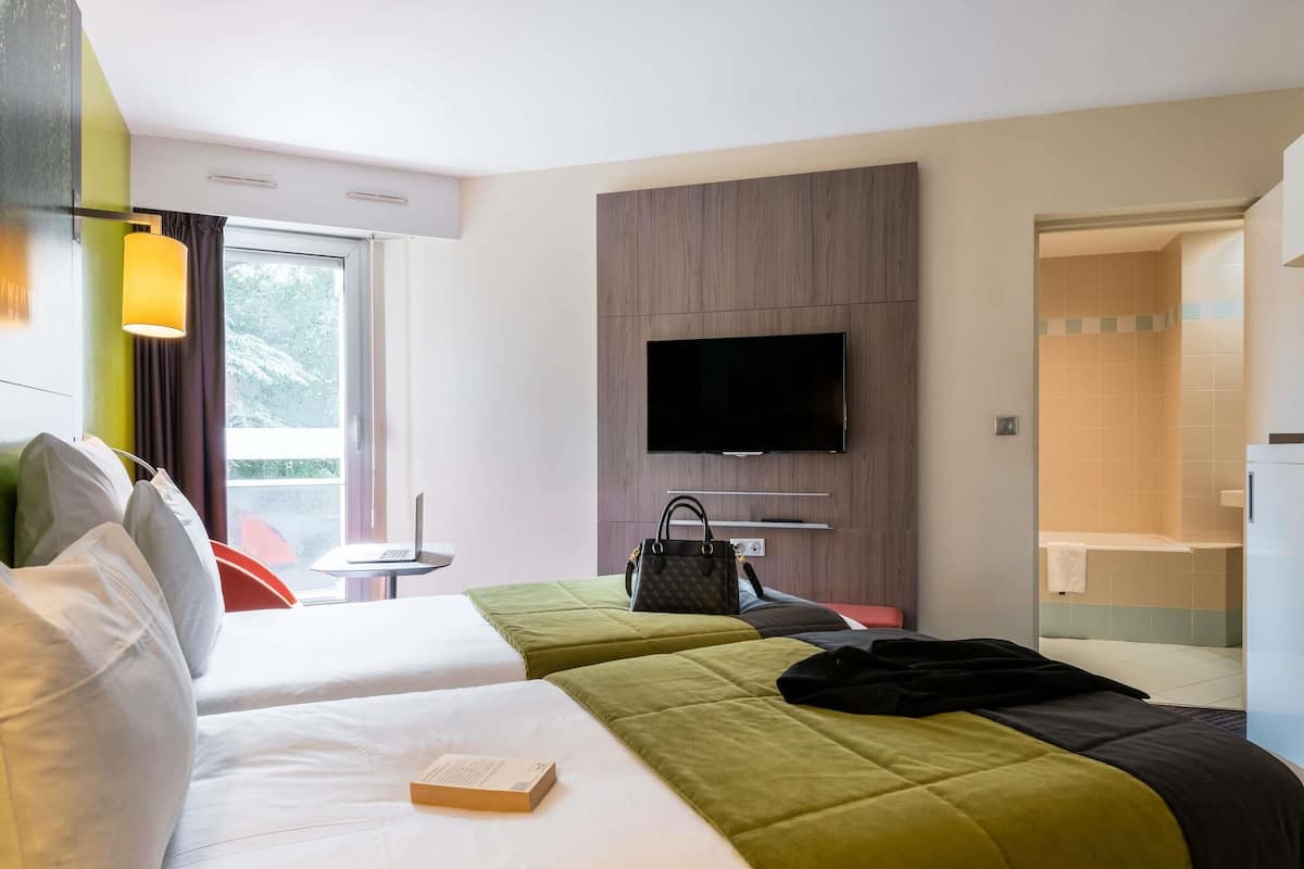 A hotel room with two beds, a TV mounted on the wall, a small table with a laptop, and a view of the outdoors through a window.