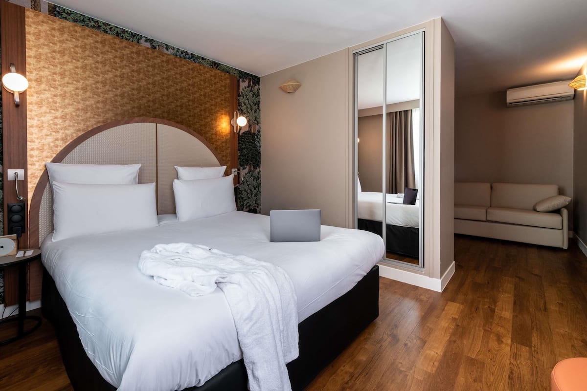 A modern hotel room with a large bed, a laptop, a sofa, and a mirror.