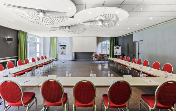 Une salle de conférence avec de longues tables, des chaises rouges et des ventilateurs de plafond.