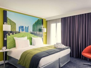 Una camera d'albergo con un letto grande, vista sulla città dalla finestra, una poltrona rossa e un quadro appeso al muro.