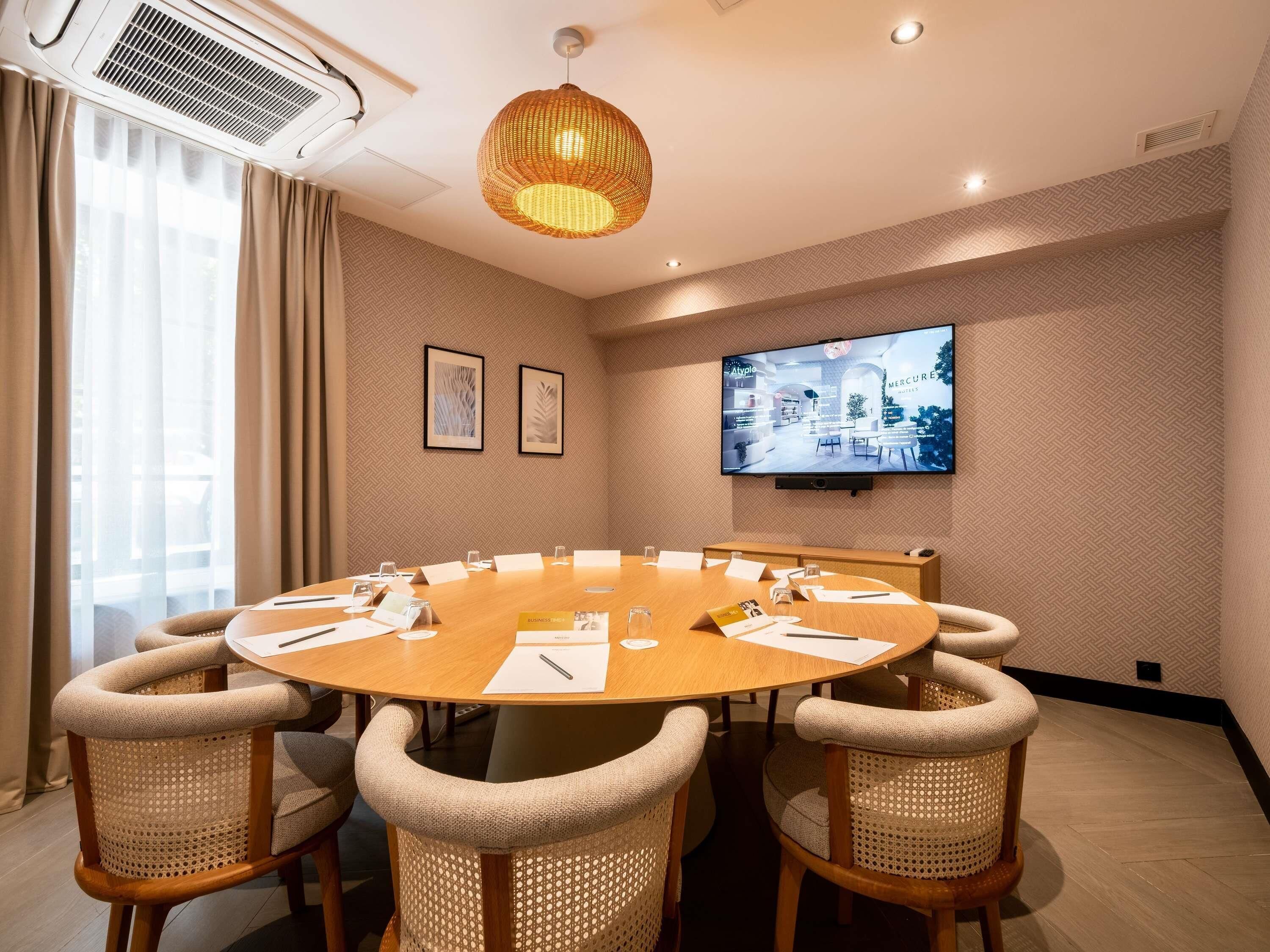 A conference room with a round table, chairs, a television screen, and a ceiling light fixture.