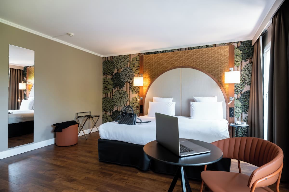A hotel room with a bed, a laptop on a small table, a chair, and a trash bin.