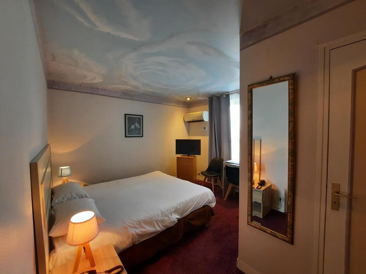 A hotel room with a bed, bedside lamp, TV, desk, chair, and a mirror on the wall.