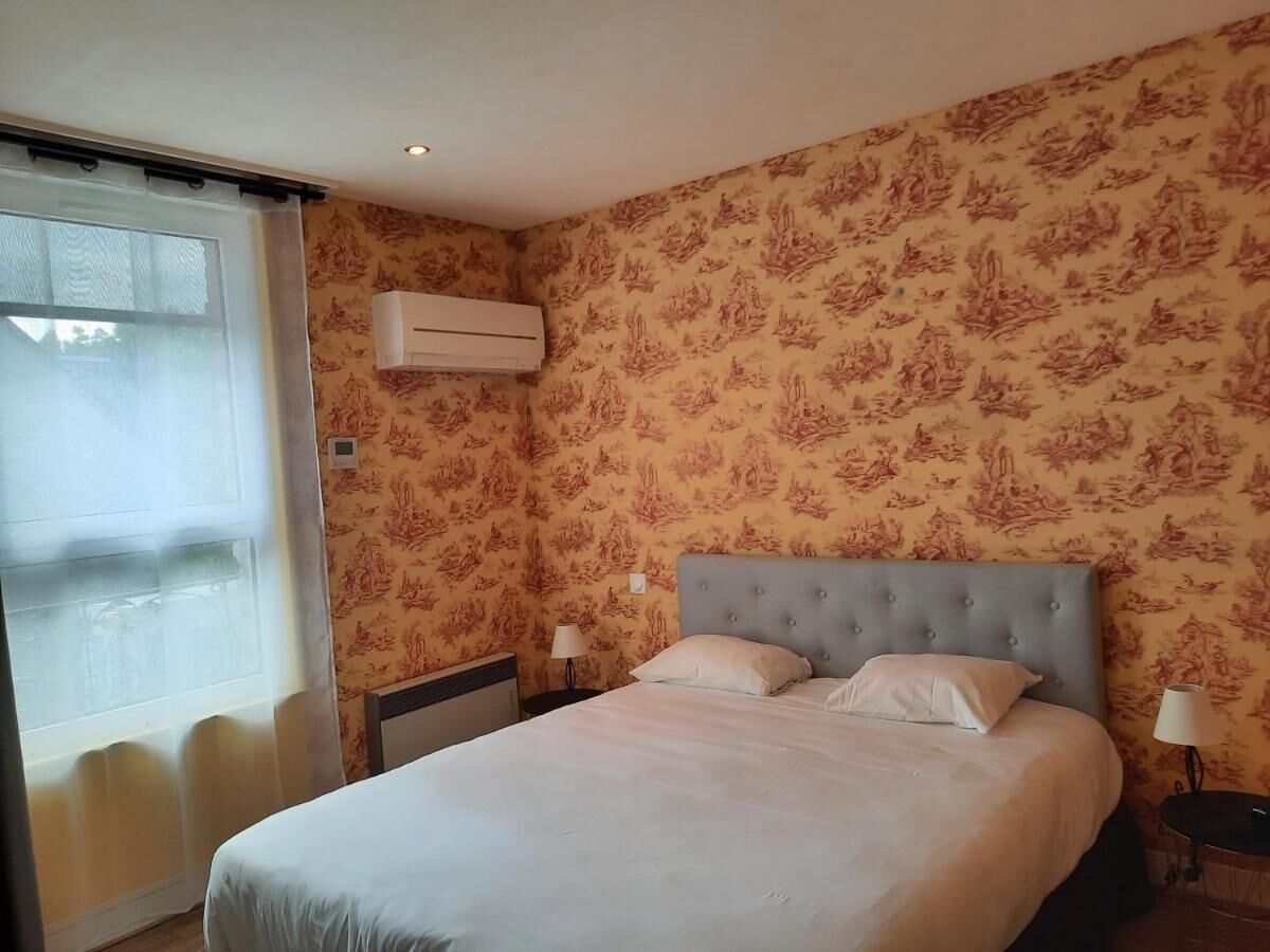 A hotel room with a bed, two bedside tables, a window with curtains, an air conditioning unit, and patterned wallpaper.
