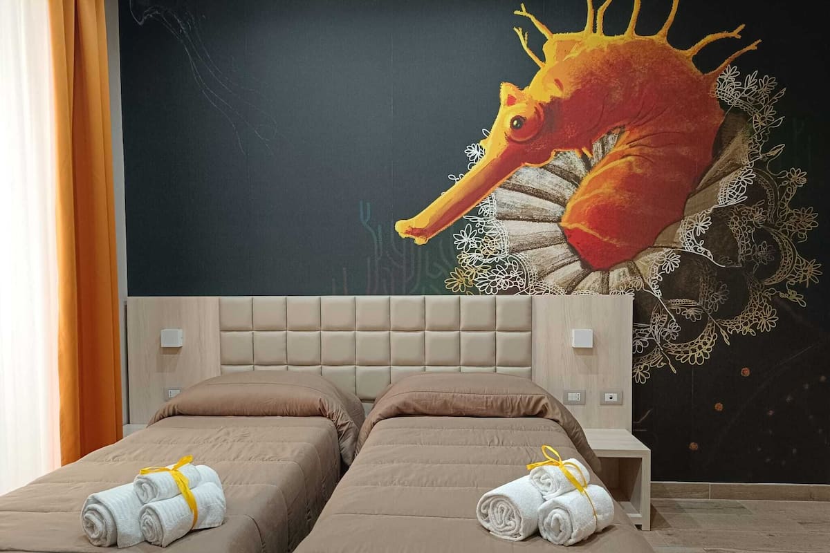 Habitación de hotel con dos camas, cada una con cabecero color beige y ropa de cama a juego. Hay toallas sobre las camas y un gran mural de caballito de mar en la pared.
