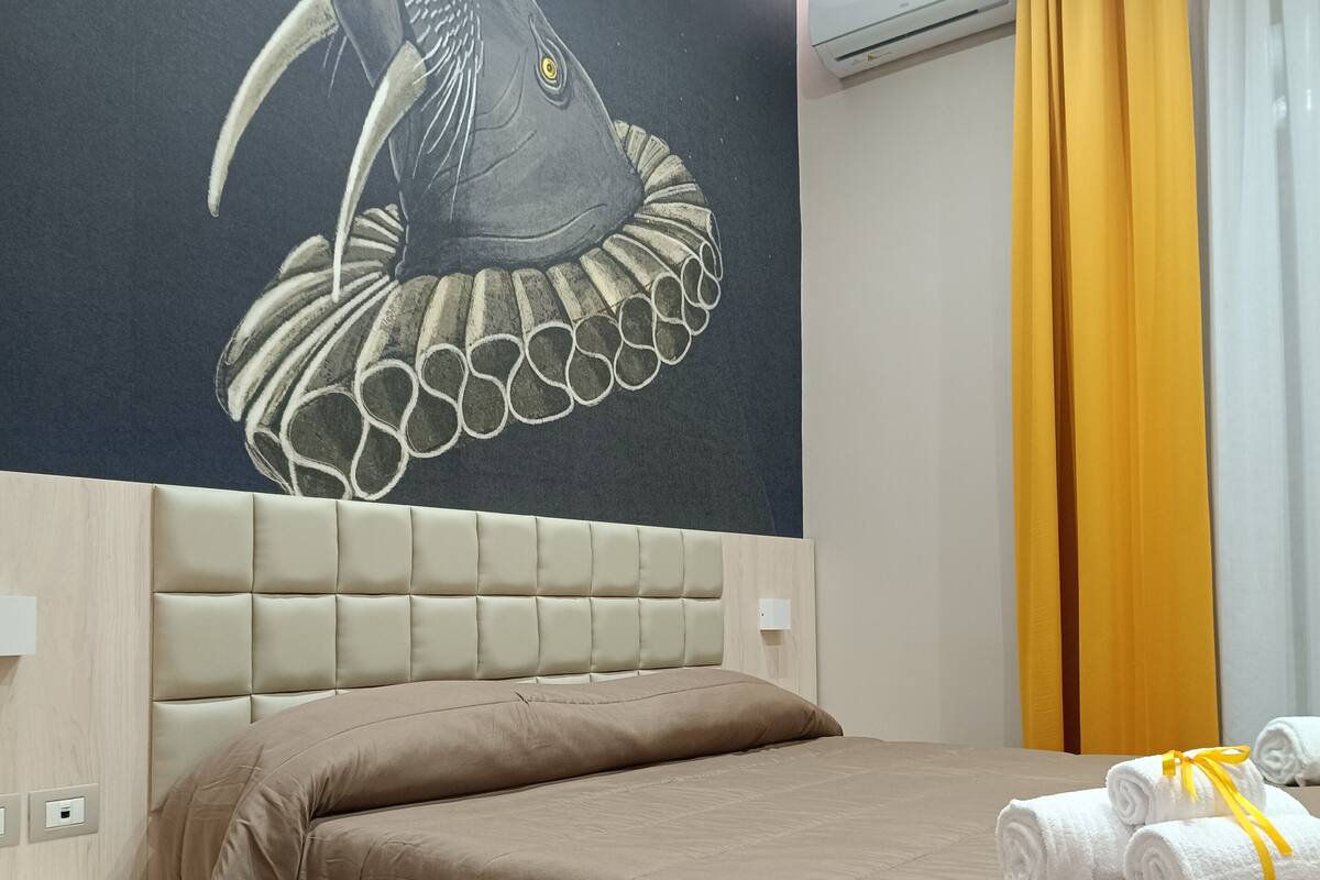Habitación de hotel con una cama grande, cabecero, un mural en la pared, una ventana con cortinas y una mesita.
