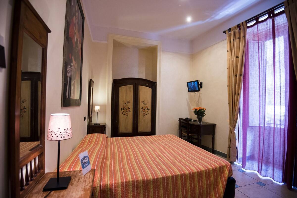 Habitación de hotel con cama, escritorio, televisión, lámpara y ventana con cortinas.
