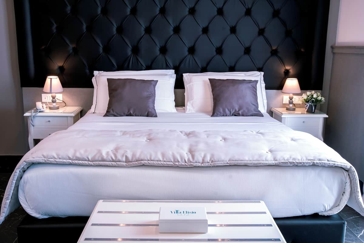 Una cama bien tendida con sábanas blancas y dos almohadas, con mesitas de noche a cada lado con lámparas y un pequeño jarrón con flores.