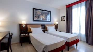 Une chambre d’hôtel avec deux lits, un bureau et une chaise. Il y a un tableau au mur et une fenêtre avec des rideaux.