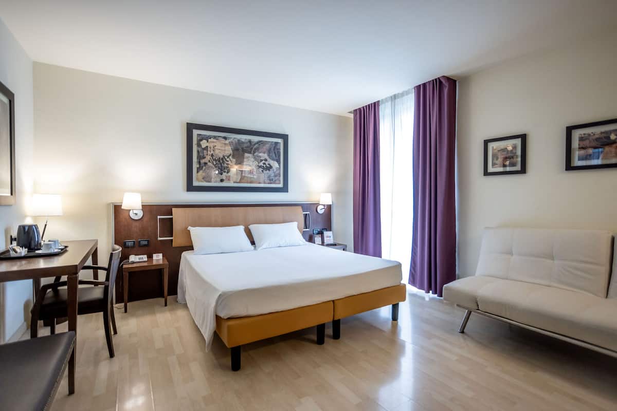 A hotel room with a bed, a desk with a chair, a sofa, and two framed pictures on the wall.
