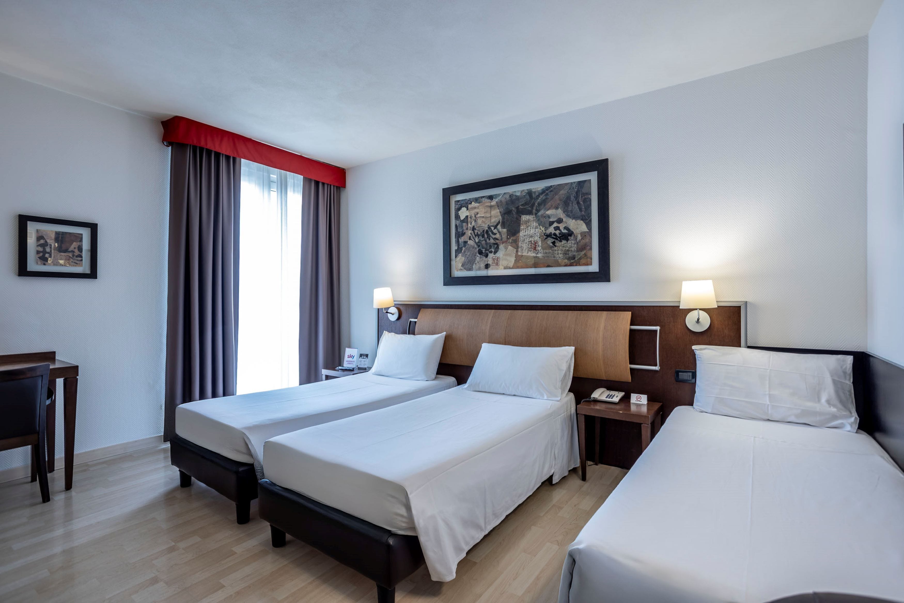 A hotel room with two beds, a desk, a chair, a nightstand with a lamp, and a framed artwork on the wall.