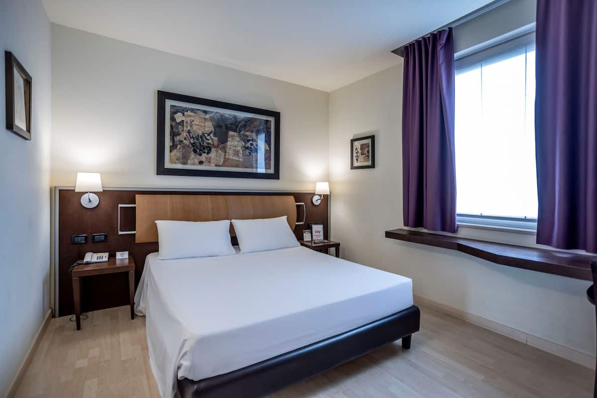 A hotel room with a large bed, wooden headboard, two bedside tables with lamps, a framed artwork above the bed, and a window with curtains.