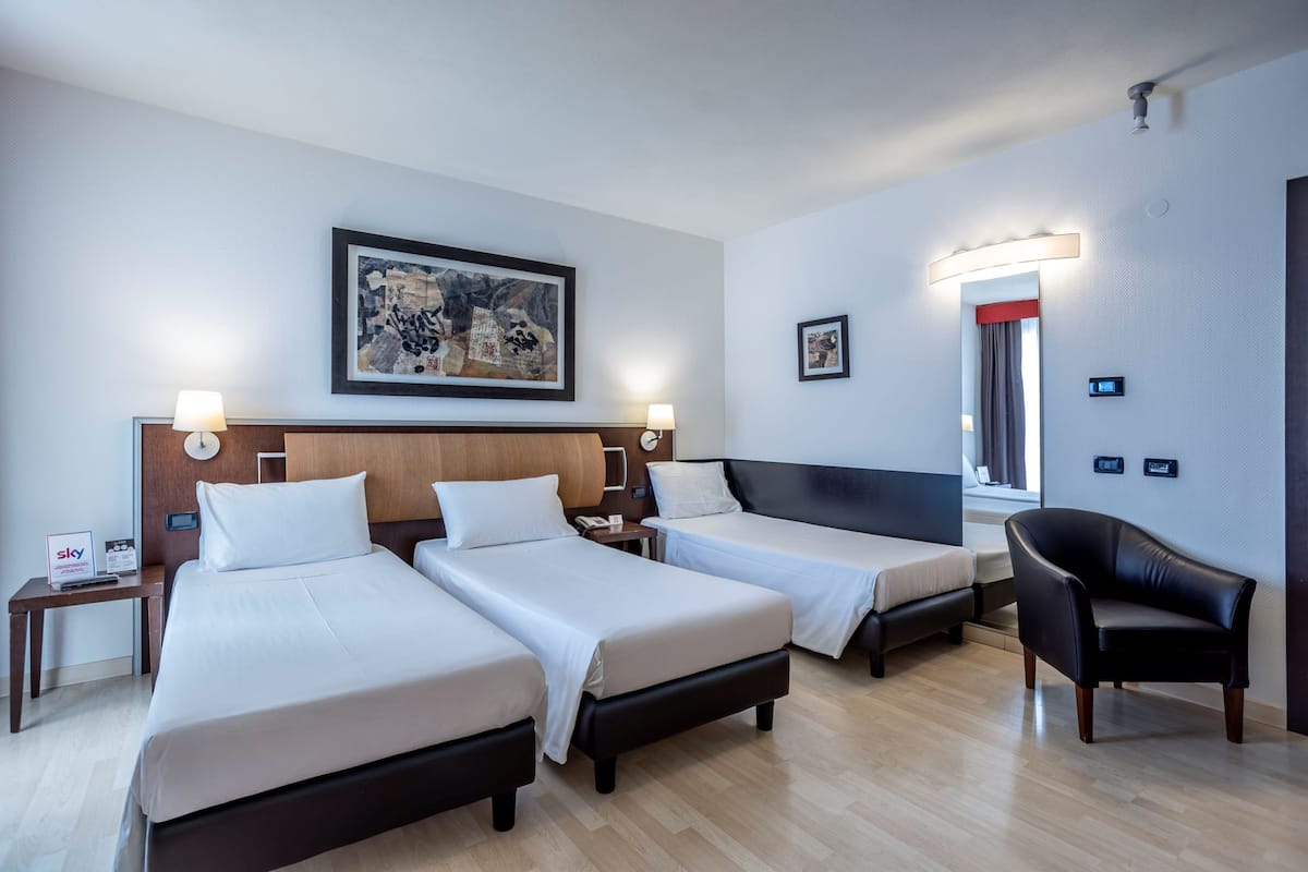 A hotel room with three beds, a desk, a chair, and a painting on the wall.