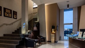 A modern lobby with a staircase, seating area, reception desk, and large windows offering a view. - Hotel Stadio (Piacenza)