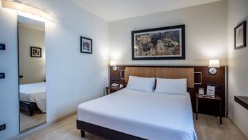Habitación de hotel con una cama grande, cabecera de madera, mesita de noche con lámpara, un cuadro enmarcado en la pared y un espejo en la puerta.