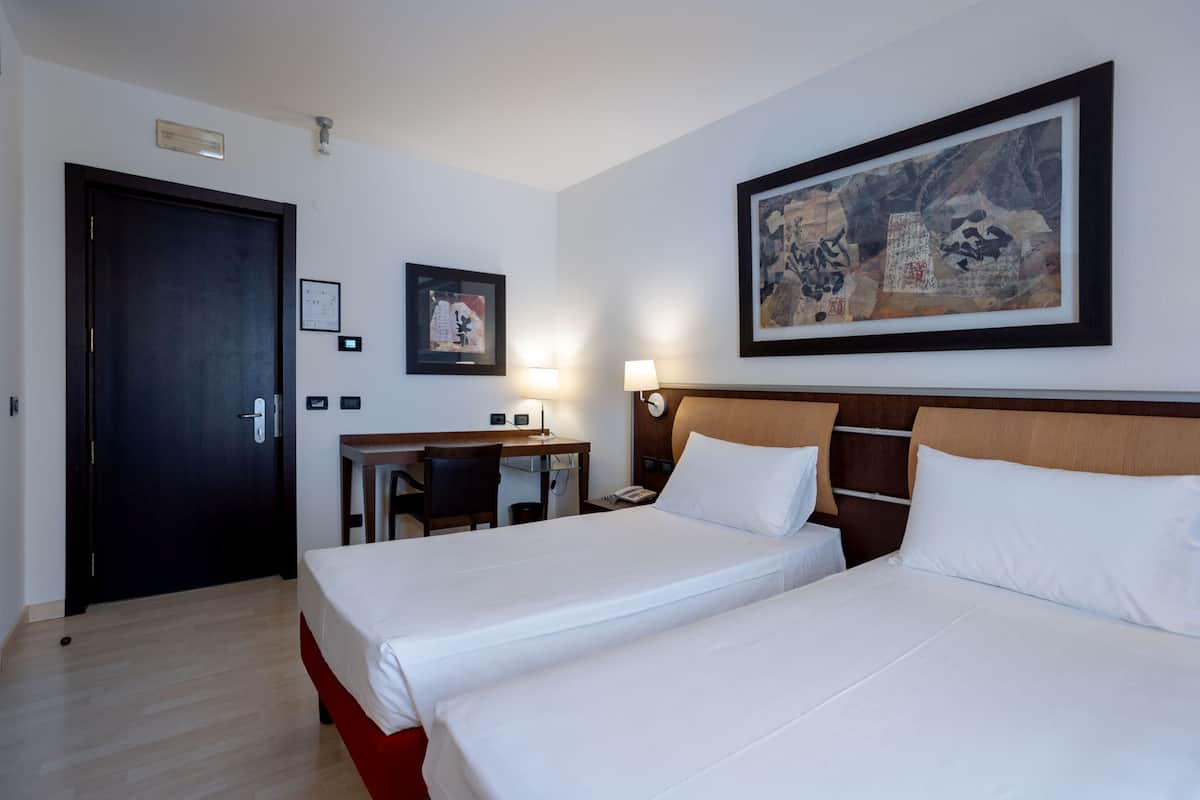 A hotel room with two beds, a desk, a chair, a phone, and a framed artwork on the wall.