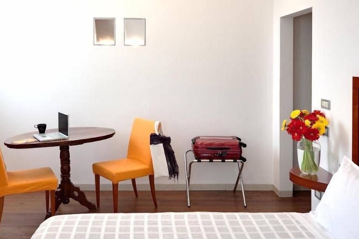 Habitación de hotel con una cama, una mesa redonda de madera con una computadora portátil, dos sillas, una maleta y un florero con flores.