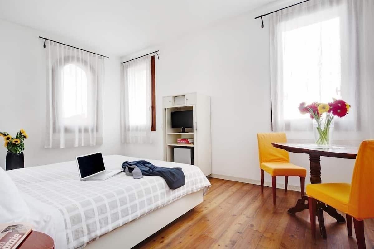 Un dormitorio con cama, escritorio, silla, ordenador portátil, estantería y ventana con cortinas.