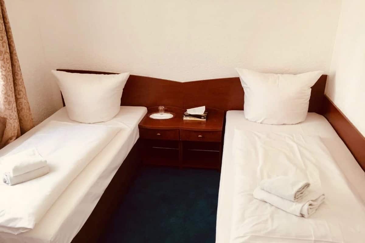 Een hotelkamer met twee enkele bedden, elk voorzien van een kussen en een opgevouwen handdoek. Tussen de bedden staat een houten nachtkastje met een lamp en een bord.
